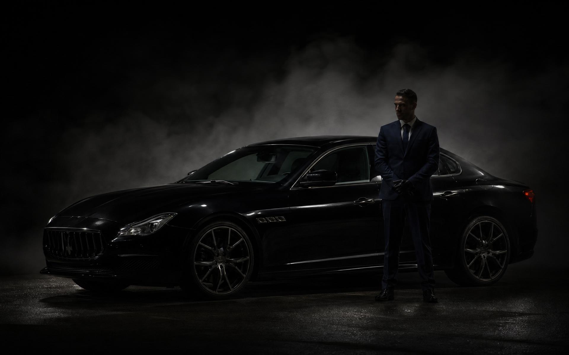 Chauffeur in a dark suit standing beside a black Maserati